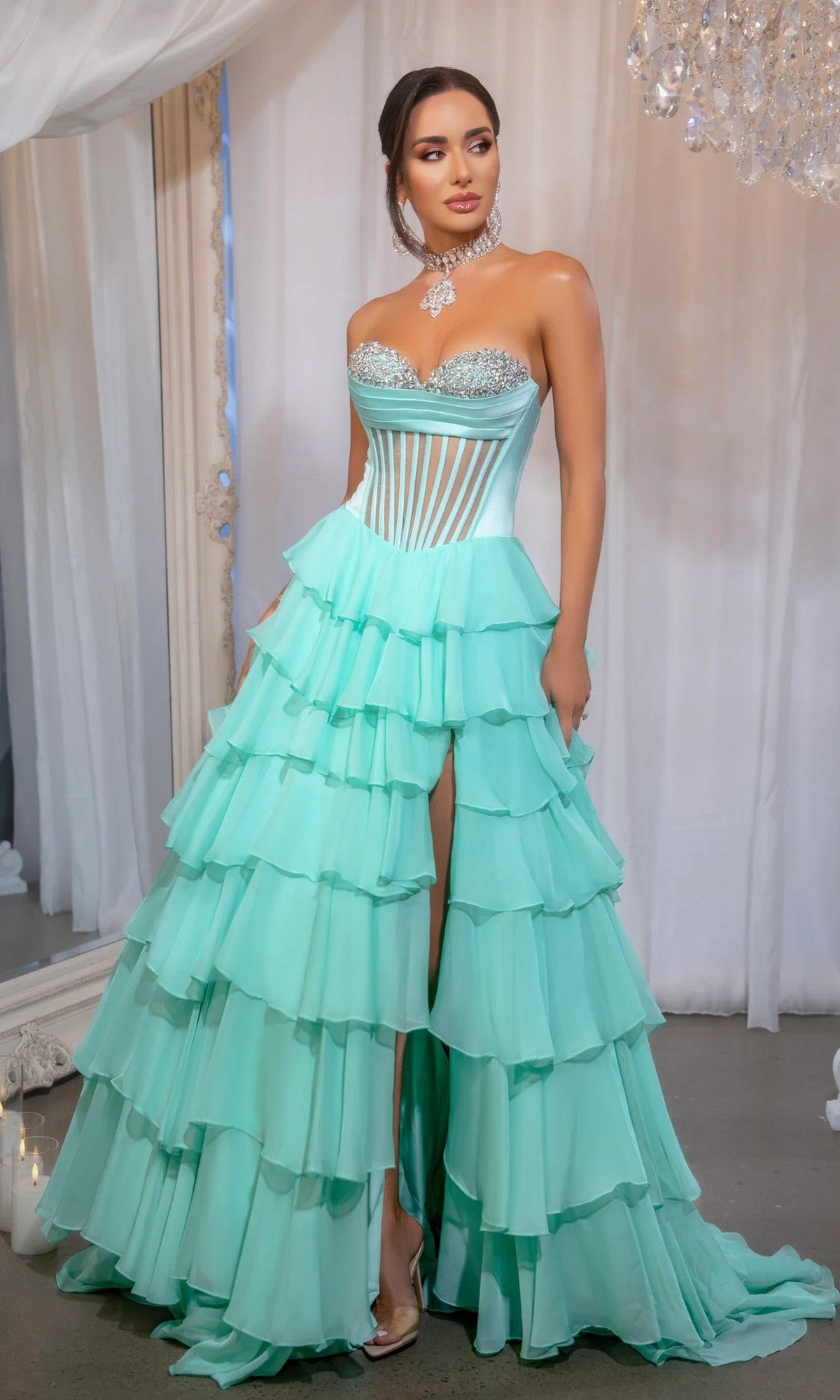 A woman stands indoors near white drapes and a chandelier, wearing the Portia and Scarlett PS25961 long mint prom dress with a strapless corset bodice, tiered ruffle skirt with high slit, and silver heels.